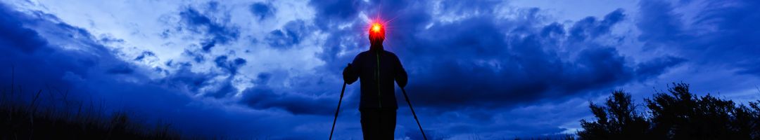 Person mit Stirnlampe und Trekkingstöcken vor dunkelblauem Himmel