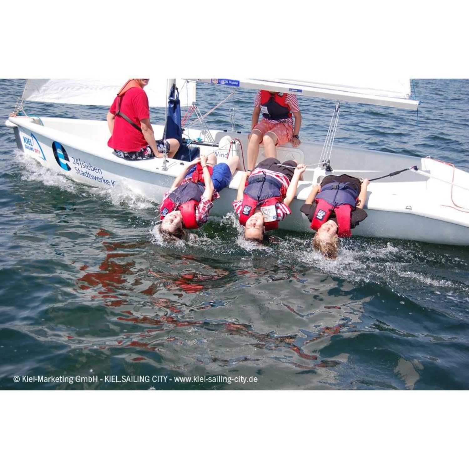 fünf Personen mit Secumar Echo Schwimmhilfe/ Kajakweste blau/ rot auf Segelboot, drei lehnen sich ins Wasser